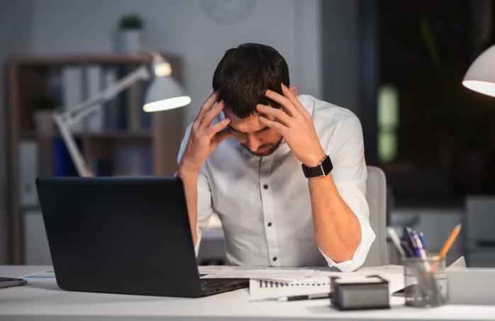 Stressed businessman reviewing financial documents at desk late at night with laptop