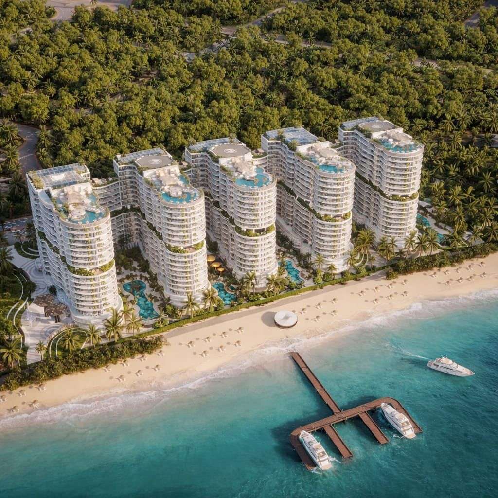 An aerial view of the AMRA by Citi Developers residential resort complex, featuring several curved, modern high-rise buildings situated on a sandy beach surrounded by palm trees, with a pier and boats in the water.