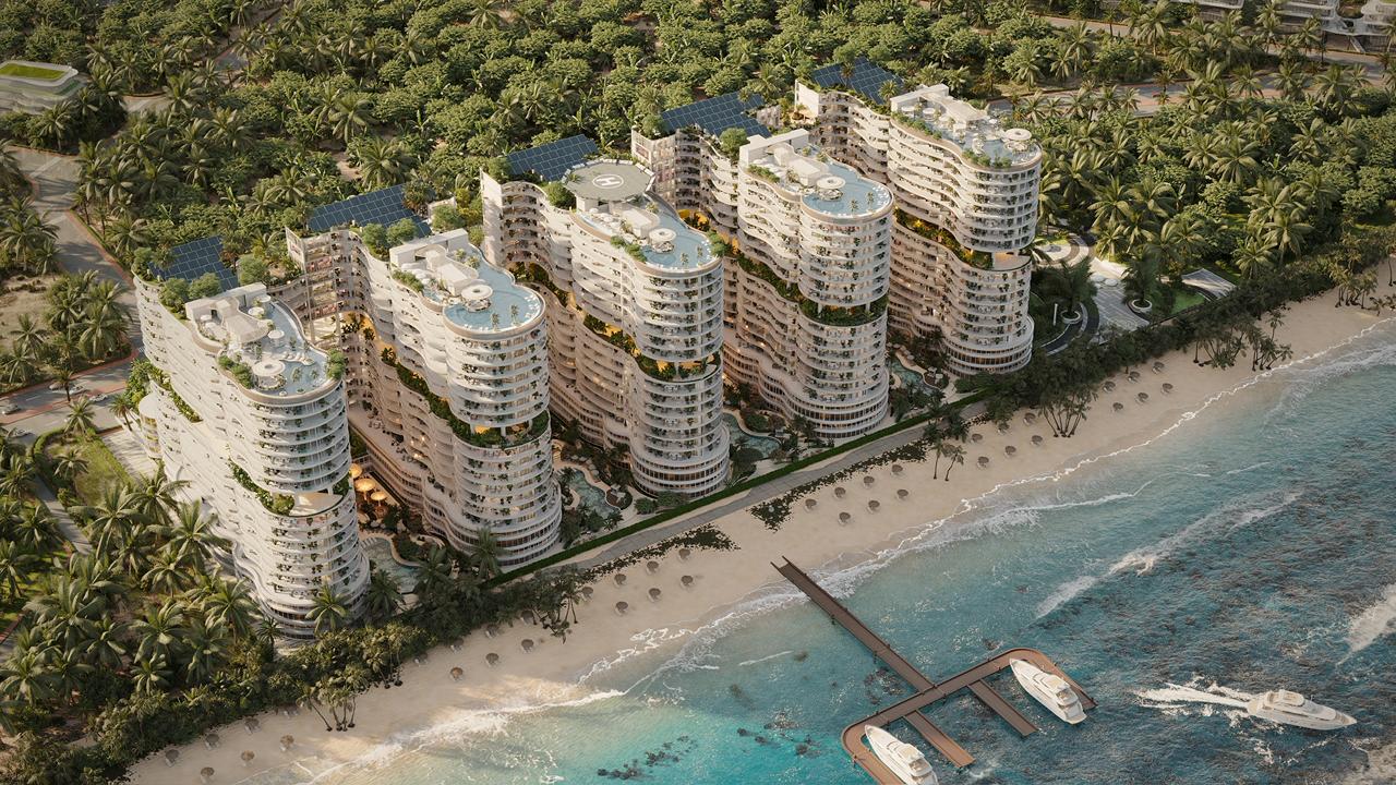 An aerial view of the AMRA by Citi Developers residential resort complex, featuring several curved, modern high-rise buildings situated on a sandy beach surrounded by palm trees, with a pier and boats in the water.